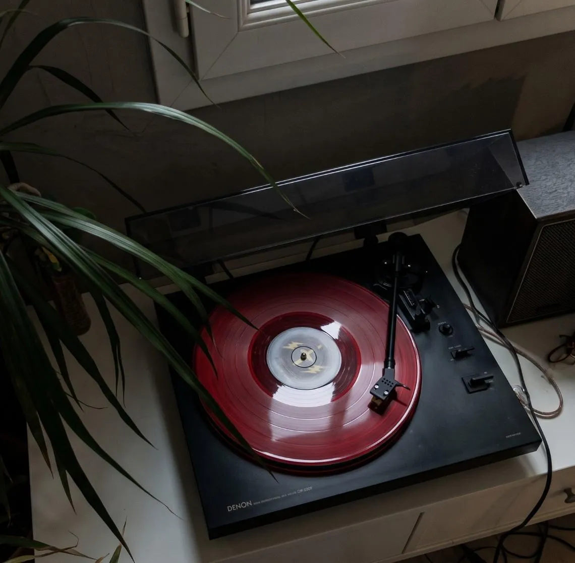 Toca-discos com um vinil vermelho tocando, ao lado de uma planta e de uma caixa de som